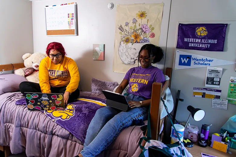 two students sitting on a bed in a dorm room working on laptops