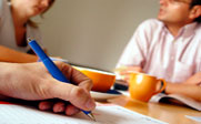 A photo of three people setting at a table with two of them talking and the other taking notes.
