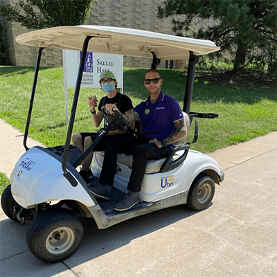 Photo of two people riding on a golf cart handing out stuff