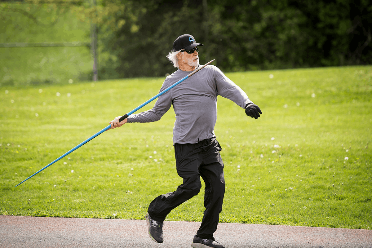 An senor man preparing to throw a javelin on a grassy field, wearing a gray shirt, black pants, and a cap.