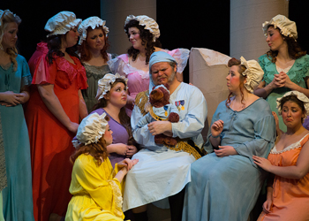 A man in a nightcap with a teddy bear is surrounded by women.