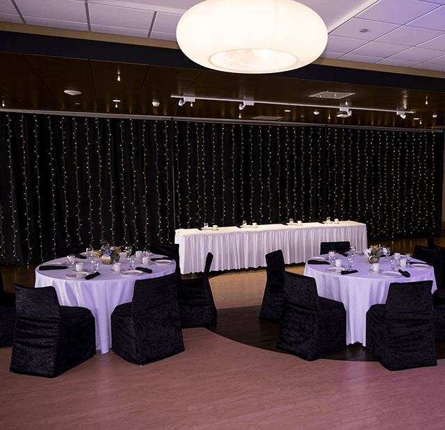 A formal dining setup with round tables in white tablecloths and black chairs, a long table with string lights in the background.