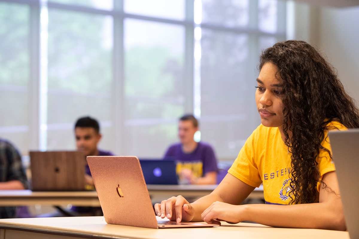 female student sitting at a table working on a laptop in a classroom setting