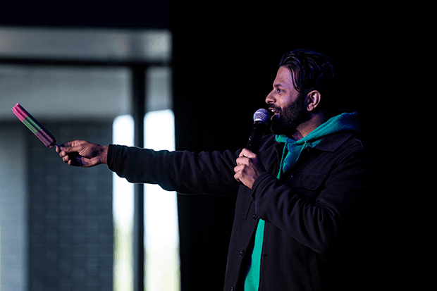 Comedian Asif Ali on stage holding a microphone to his face and a popsicle out to the audience, with a dark background and bright light from a window.