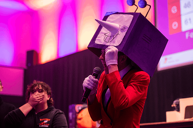 A person in a costume lifts a cartoon face box headpiece to reveal themselves, speaking into a microphone on a brightly lit stage. Another person is visible, wearing a dark hoodie and covering their mouth with their hand, seemingly amused or surprised.