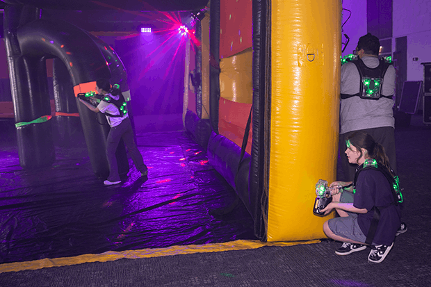 Three people playing laser tag in a dimly lit room with glowing green vests and inflatable barriers.