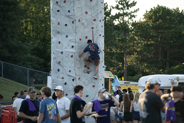 Person climbing a tall artificial rock wall at an outdoor event with people milling around.